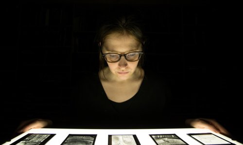 A person looks an an illuminated manuscript on a table, with dark behind them it is as if they are silhouetted with the book.
