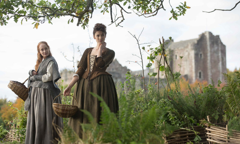 Two cast members from Outlander in a field with Doune Castle behind them